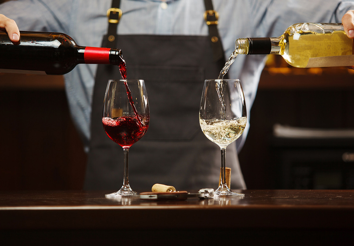 Сомелье-мужчина наливает красное и белое вино в бокалы на длинной ножке